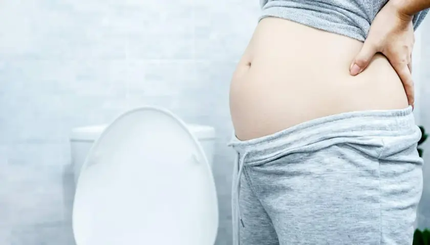 Frau mit Blähbauch hält sich den Bauch. Was hilft gegen Blähbauch? Die Toilette im Hintergrund deutet auf Verdauungsprobleme hin.