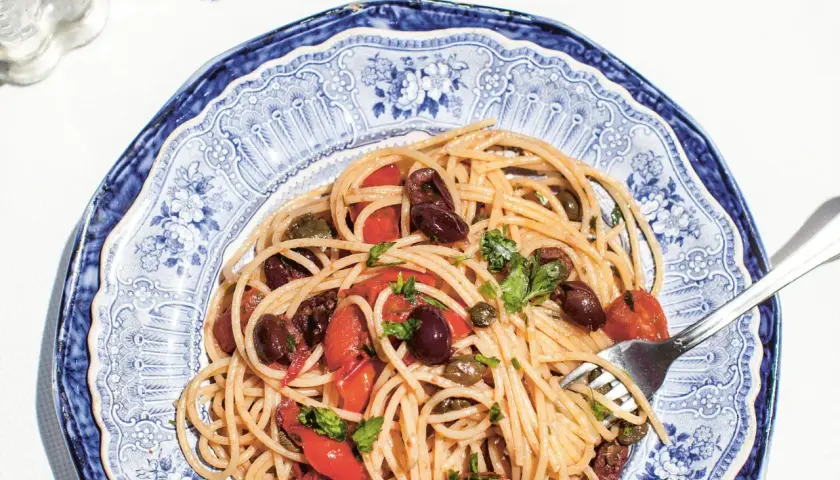 Spaghetti Puttanesca mit Oliven, Kapern und Tomaten auf einem blau-weißen Teller.