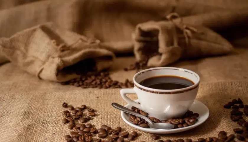 Aromatic coffee beans spill from burlap sacks, surrounding a white cup of dark brew. Zastanawiasz się, ile kofeiny ma kawa parzona?