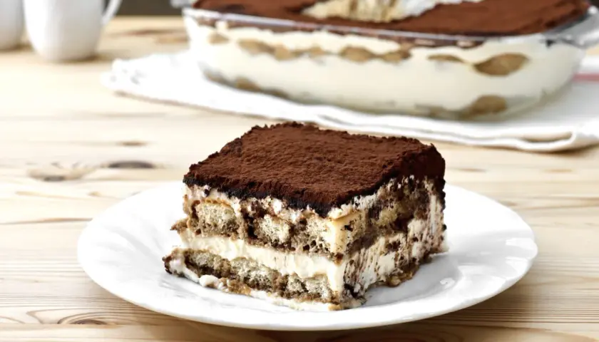 Una fetta di tiramisù su un piatto bianco, con la teglia sullo sfondo. La crema tiramisù perfetta, soffice e stratificata, è cosparsa di cacao.