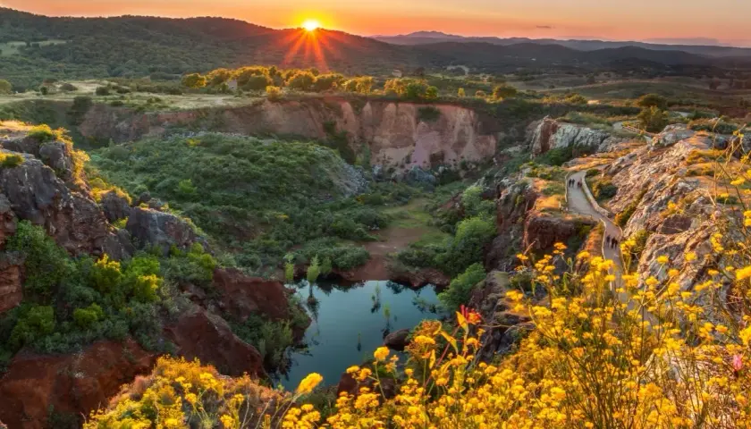 Atardecer dorado sobre un cañón con un estanque, ideal para senderismo Sevilla. Flores amarillas bordean el camino.