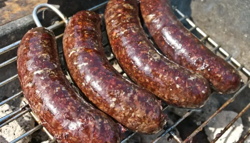 Cztery soczyste kaszanki na grilla, piekące się na ruszcie. Dym unosi się, zapowiadając pyszny smak.