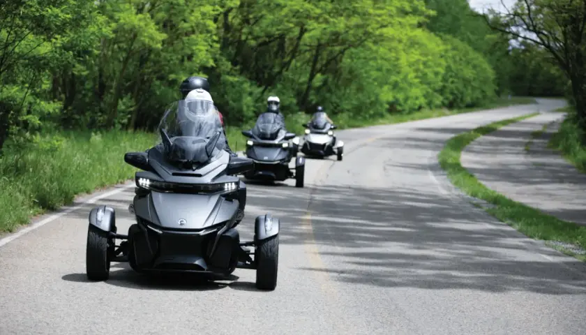Trzy pojazdy typu Can-Am Spyder jadące po krętej drodze wśród zieleni. Ich nowoczesny design i dynamiczna jazda tworzą wrażenie efektu shimmy.