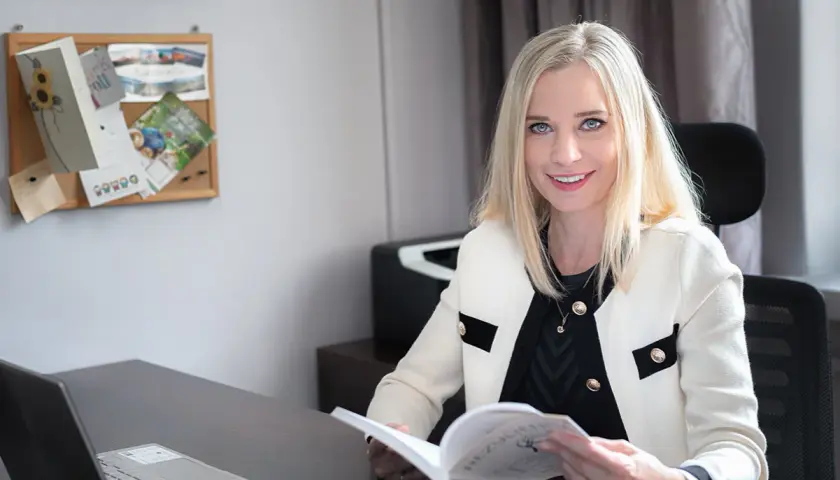 Uśmiechnięta blondynka w białej marynarce siedzi przy biurku i czyta książkę.