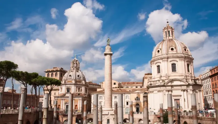 La colonna di Traiano si erge maestosa a Roma, con cupole barocche e pini marittimi sotto un cielo azzurro e nuvoloso.