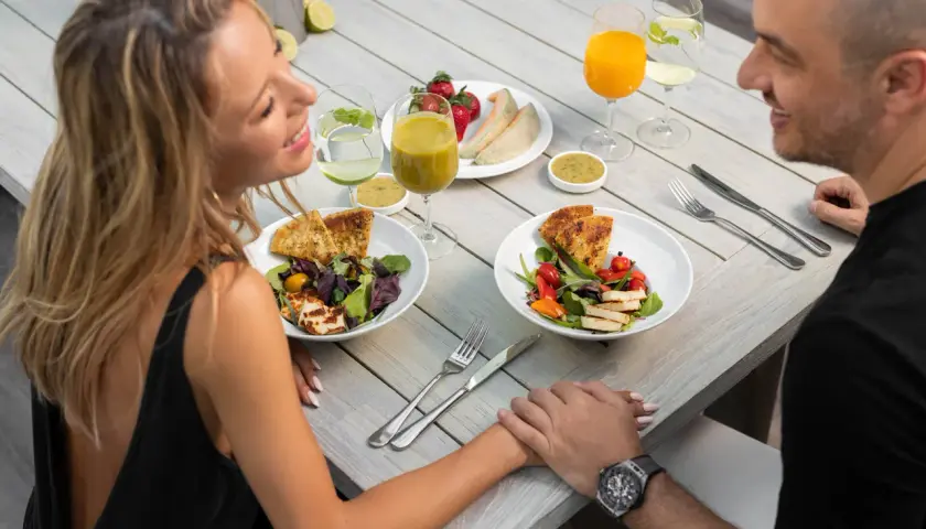 Romantyczny catering dla par. Uśmiechnięta para trzyma się za ręce przy stole z pysznymi sałatkami, owocami i napojami.