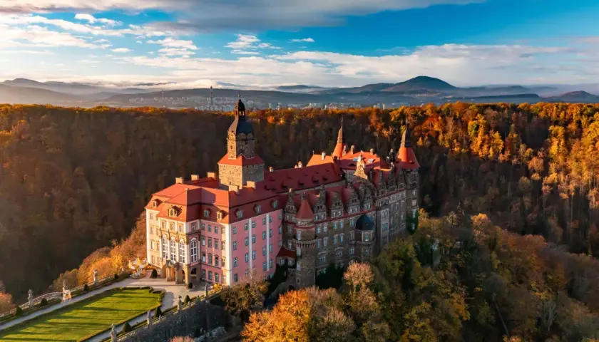 Malowniczy hotel zamek otoczony jesiennym lasem, z wieżą i czerwonymi dachami, na tle gór i błękitnego nieba.