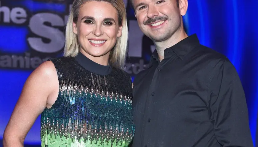 Uśmiechnięci Sławomir i Kajra pozują do zdjęcia na tle niebieskiej kurtyny. Kajra ma na sobie błyszczącą sukienkę, a Sławomir czarną koszulę.