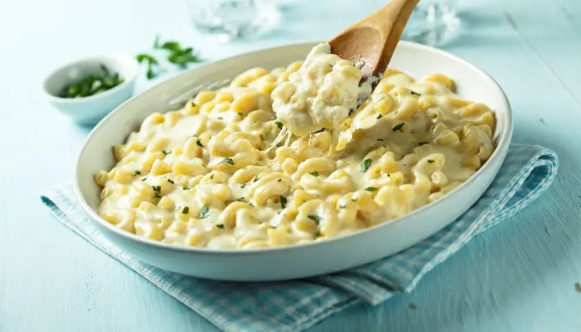 Kremowe mac n cheese z ziołami, które rozpływa się w ustach. Drewniana łyżka podnosi porcję, ukazując ciągnący się ser.