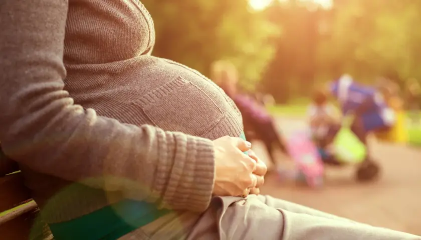 Gravid kvinna sitter på en parkbänk, njuter av solen. Hon håller sin mage, som om hon väntar på sitt barn. I bakgrunden syns andra människor och barnvagnar. Det är en varm och solig dag, med gula flytningar i luften.