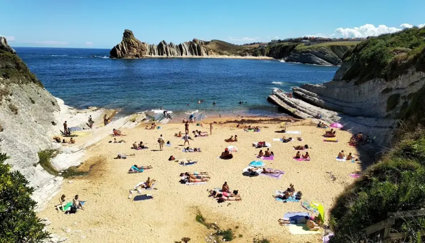 Gente disfrutando del sol y el mar en la playa de cerrias, con formaciones rocosas impresionantes.