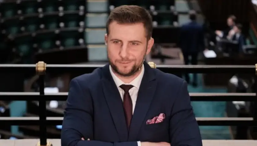 Mężczyzna w garniturze z krawatem i poszetką, siedzi na tle sali sejmowej.