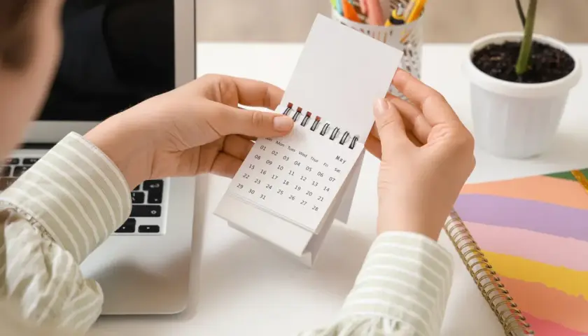 Ręce przeglądają kalendarz, szukając lipiec święta wolne od pracy. Obok laptop i kolorowy notatnik.