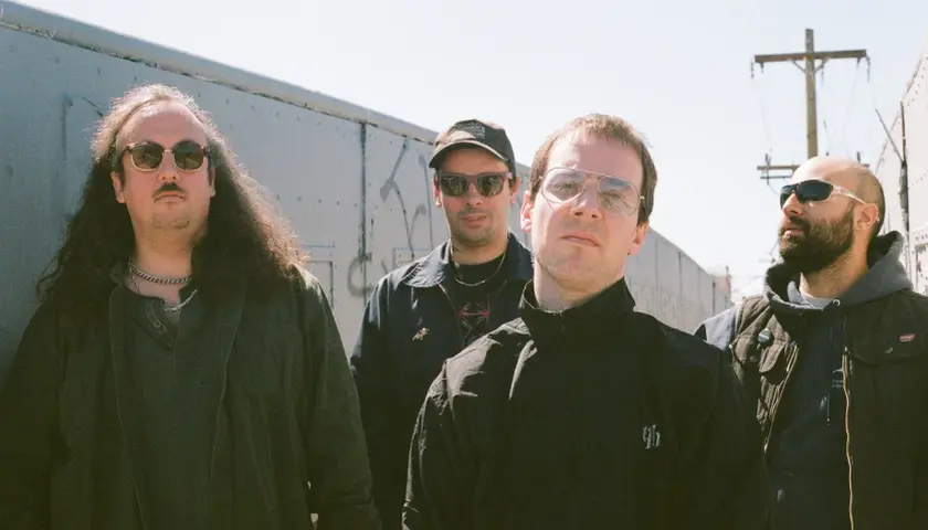 Czterech mężczyzn, jakby z zespołu The Sisters of Mercy, pozuje na tle wagonów kolejowych.