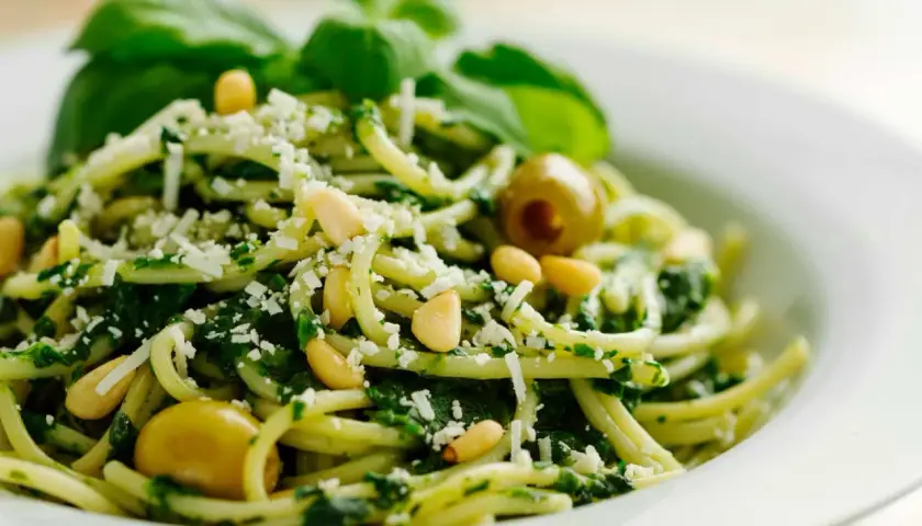Pasta con pesto de pistacho, piñones, aceitunas verdes y queso rallado, decorada con hojas de albahaca fresca.