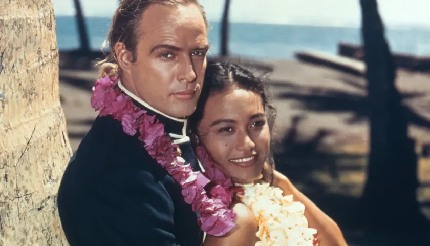Marlon Brando z kobietą w kwiatowych girlandach na tle palmy i oceanu.