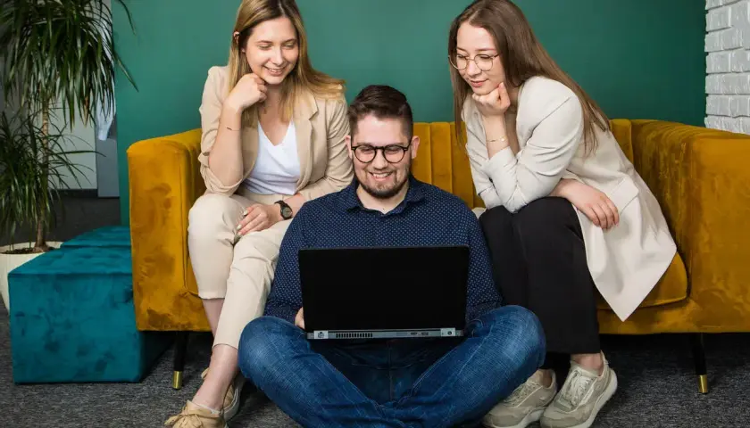 Trzy osoby pracują przy laptopie. Dwie kobiety siedzą na kanapie, a mężczyzna na podłodze. Zastanawiają się, cro co to jest.