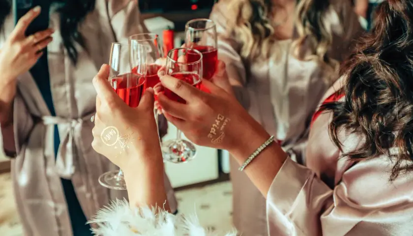 Dłonie w szlafroczkach unoszą kieliszki z czerwonym napojem, na jednej widoczny tatuaż "Bride".