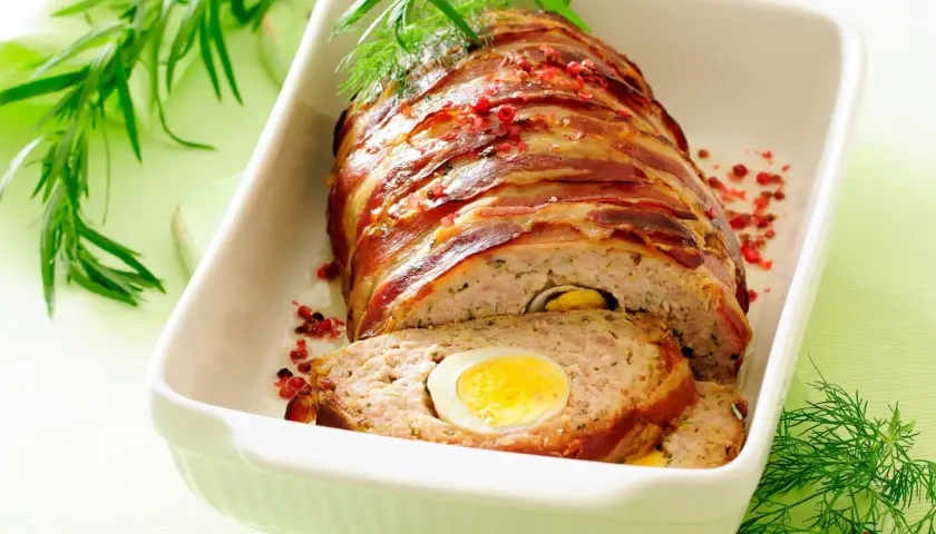 Najlepsza pieczeń rzymska, zawinięta w boczek, z jajkiem w środku, podana w białej brytfannie, ozdobiona ziołami i pieprzem.
