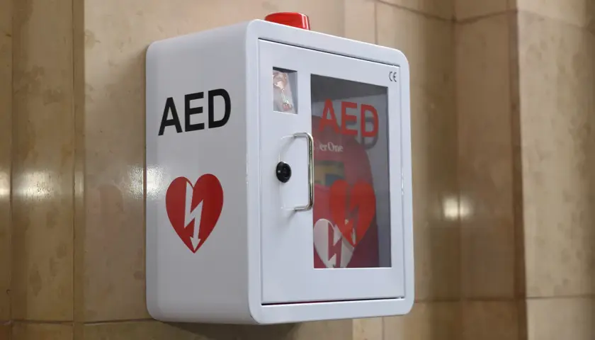 Biała obudowa AED z czerwonym sercem i symbolem błyskawicy, zamontowana na ścianie.
