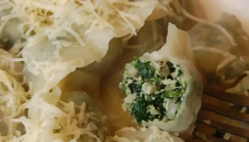 Pyszne pierogi ze szpinakiem, polane sosem i posypane startym serem. Jeden pieróg jest przekrojony, ukazując soczyste nadzienie.