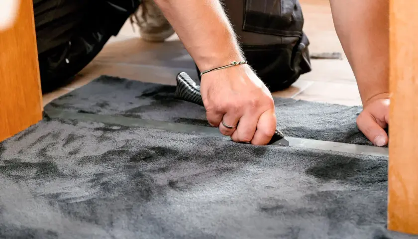 Mann schneidet mit einem Teppichmesser einen grauen Teppich zu. Das Verlegen des Teppichs wird präzise ausgeführt.