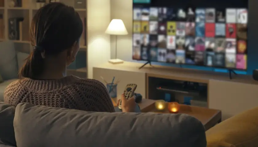 Kobieta z pilotem w ręku ogląda film na telewizorze, zastanawiając się, jak podłączyć komputer do telewizora, by cieszyć się większym ekranem.