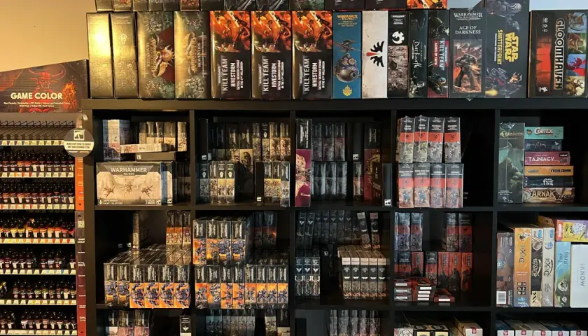 Półki pełne pudełek z grami i figurkami, w tym wiele z serii Warhammer. Widać tu zapowiedź przygód Gotreka i Felixa, a także inne tytuły.