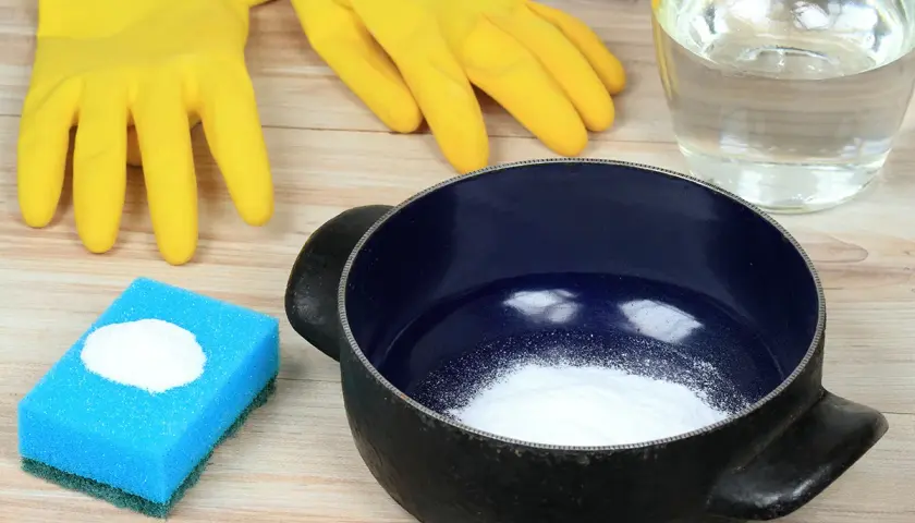 Gants jaunes, éponge bleue avec poudre blanche, casserole noire et un bocal rempli de liquide. Idéal pour savoir comment enlever le cramer dans une casserole sans bicarbonate.