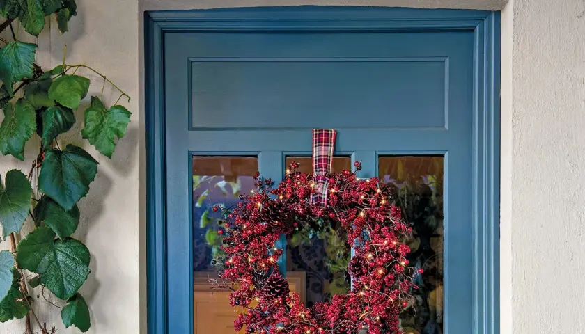 Corona navideña roja con luces y piñas colgada en una puerta azul.