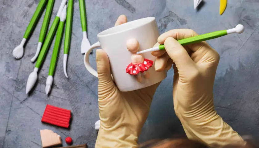 Mani guantate creano una bambolina con pasta fimo su una tazza. Tanti strumenti e colori per idee creative.