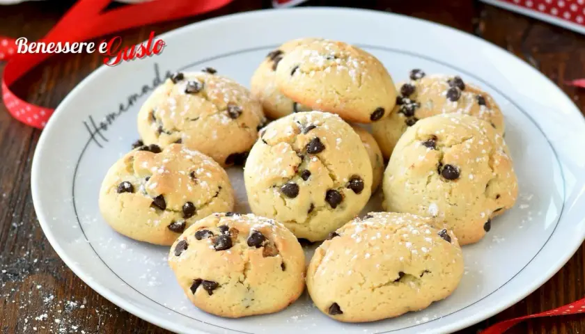 Biscotti con gocce di cioccolato spolverati di zucchero a velo su un piatto bianco, pronti per essere gustati.