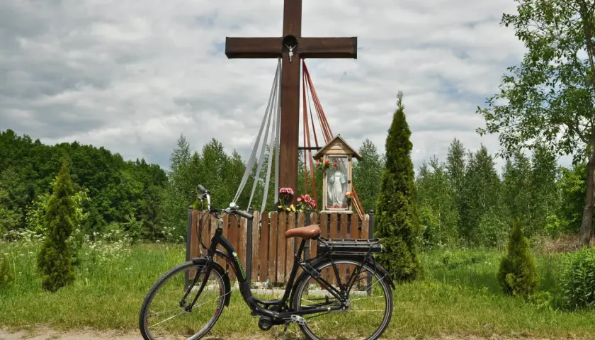 Rower przy kapliczce w Hajnówce, Białowieża. Drewniany krzyż z figurką Chrystusa i Matki Boskiej, otoczony zielenią i polnymi kwiatami.