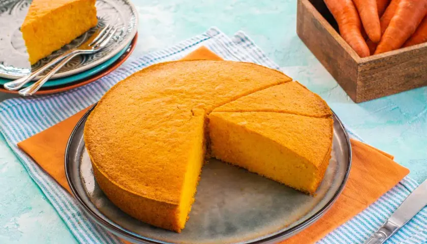 Una deliziosa torta alle carote, soffice e dorata, tagliata a fette su un piatto. Accanto, carote fresche in un cesto di legno.