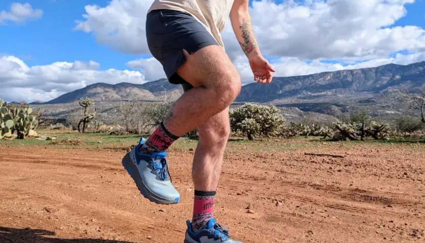 Runner's legs in Altra Timp 6 shoes and patterned socks, mid-stride on a dusty desert trail with mountains in the background.