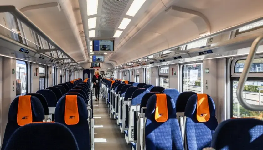 Wnętrze wagonu kolejowego z rzędami niebieskich siedzeń z pomarańczowymi zagłówkami.