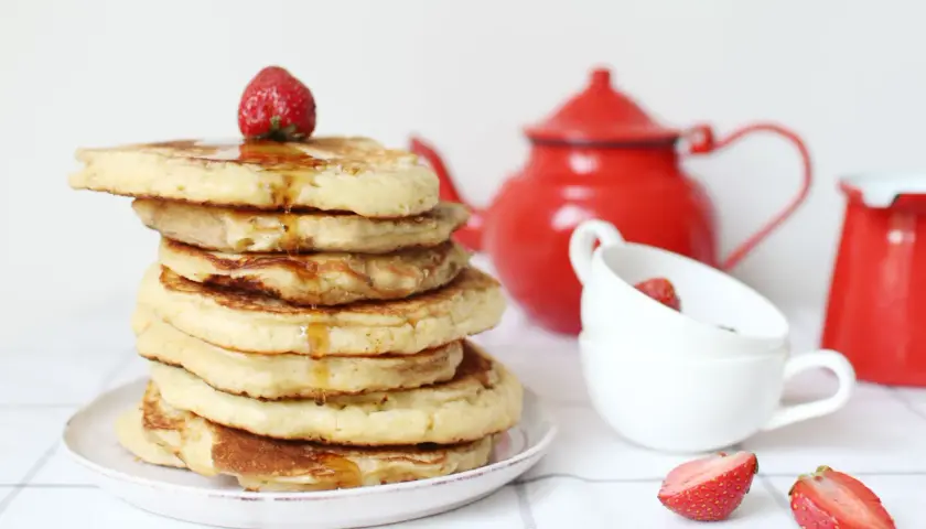 Pile de pancake sans gluten, nappés de sirop et garnis d'une fraise. Une théière rouge et des tasses complètent la scène.