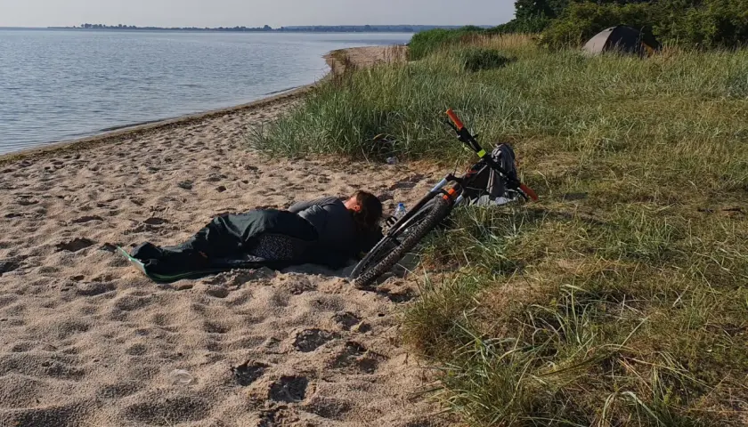 Osoba śpi na plaży obok roweru. W oddali widać wodę i drzewa. To dowód, że można spać na plaży.