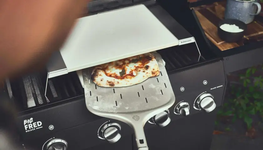 Frisch gebackene Pizza wird mit einem Pizzaschieber aus einem Gasgrill gehoben. Perfekt für den Sommer mit dem Pizza Gasgrill.