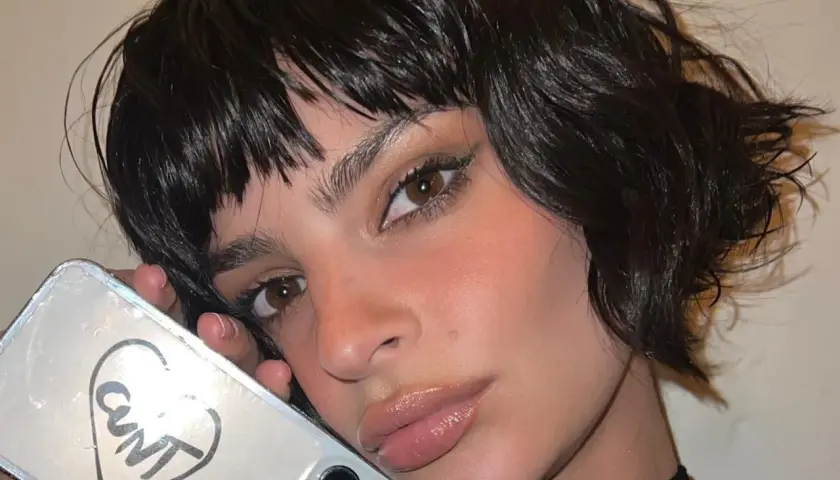 Primo piano di una donna con capelli scuri e umidi che le incorniciano il viso. Tiene in mano un telefono con un disegno a cuore e la scritta "CUNT".