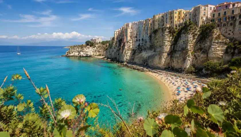A vibrant beach scene on the Calabria coast, with colorful buildings perched on dramatic cliffs overlooking turquoise waters.