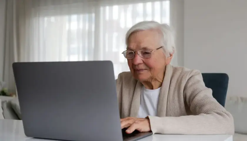 Uśmiechnięta starsza pani z okularami pracuje na laptopie, planując swoją emeryturę bez podatku.