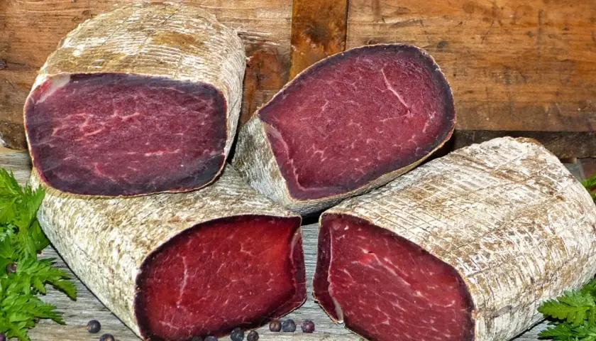 Quatre morceaux de viande des Grisons, d'origine suisse, sont présentés sur un fond de bois. La viande est rouge foncé, avec une texture fibreuse.