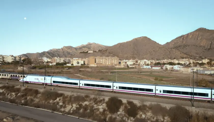 Tren AVE de Renfe, un moderno tren blanco con detalles morados, atraviesa un paisaje rural con montañas y edificios al fondo.