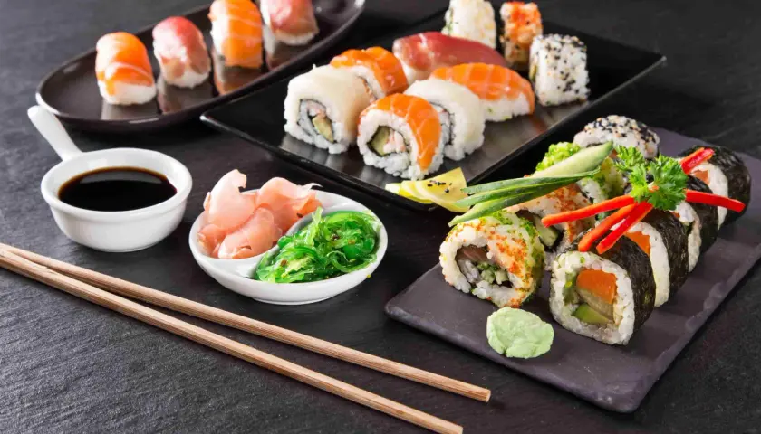 Różnorodne sushi, sos sojowy, marynowany imbir, sałatka wakame i pałeczki na ciemnym tle.
