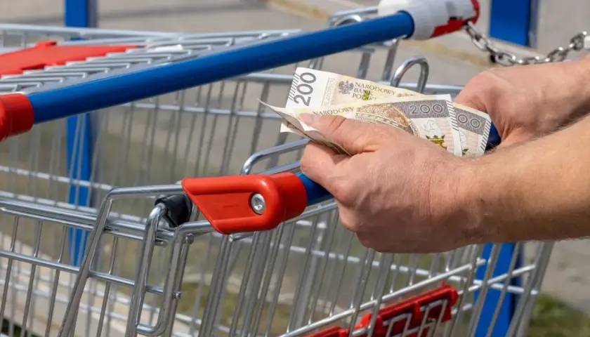 Ręce trzymają banknoty 200 zł przy wózku sklepowym. Kto zyskuje na inflacji, gdy ceny rosną?