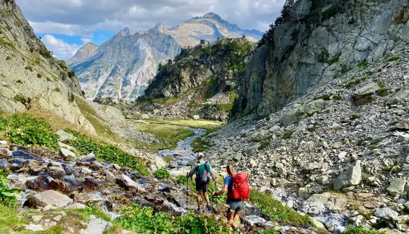 Hikers ascend a rocky path alongside a stream, with majestic mountains in the distance, on the Haute Route Pyrenees.