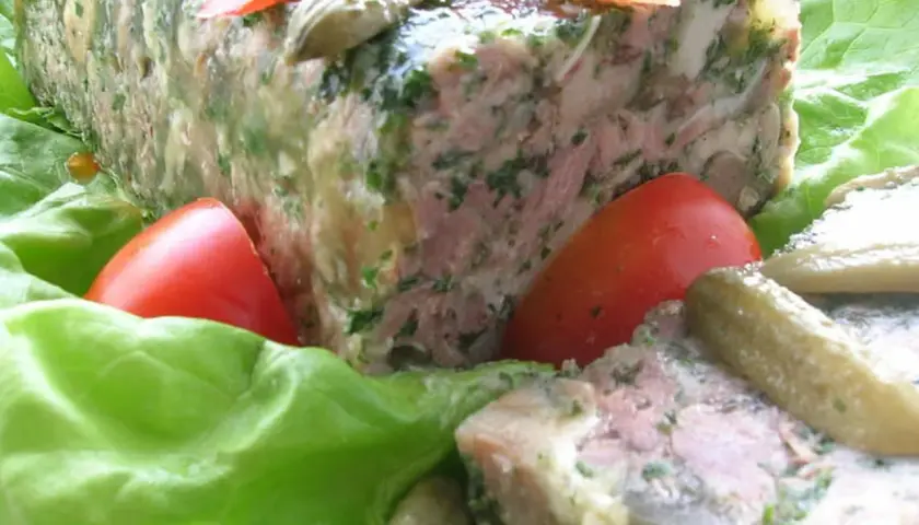 Tranche de pâté de tête maison, garnie de persil, servie sur des feuilles de laitue avec des tomates et des cornichons. Un délice pour une recette de pâté de tête.