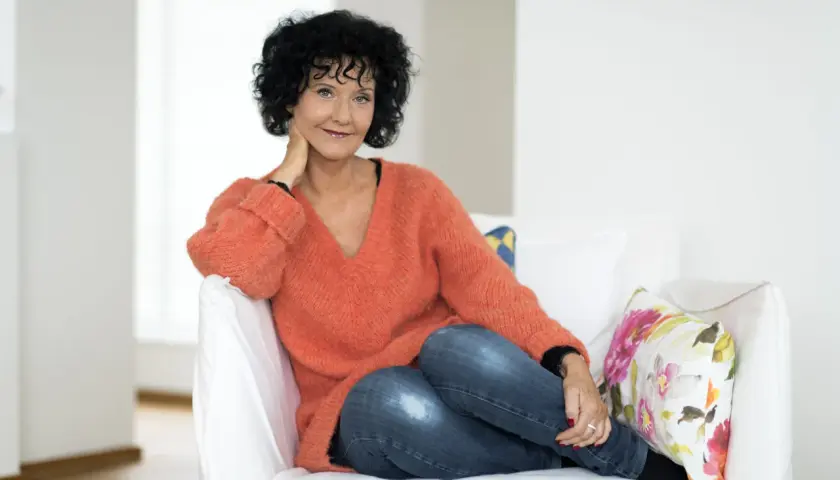 En kvinna med lockigt mörkt hår, klädd i en orange tröja och jeans, sitter avslappnat i en vit soffa. Hon ler mot kameran, med handen vid halsen. Lindell som skriver sina memoarer?