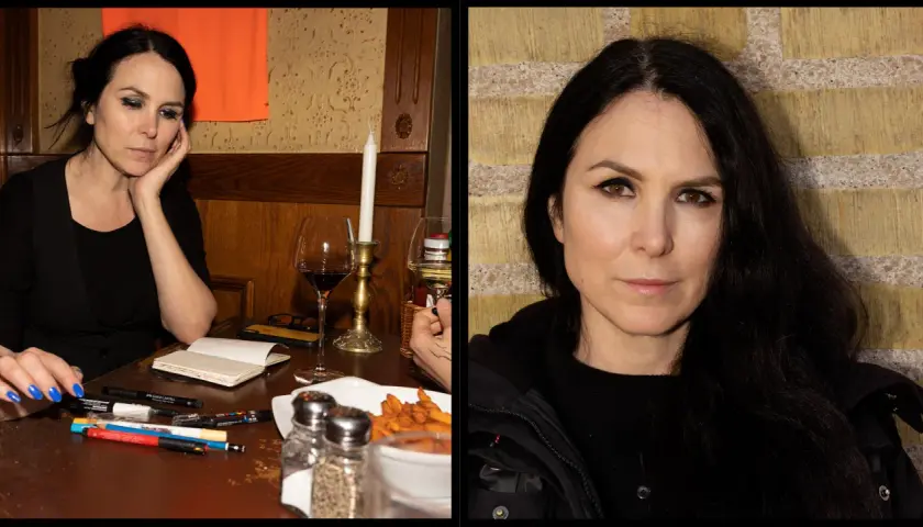 Nina Hemmingsson, med sitt karakteristiska uttryck, sitter vid ett bord med pennor och ett glas rödvin.